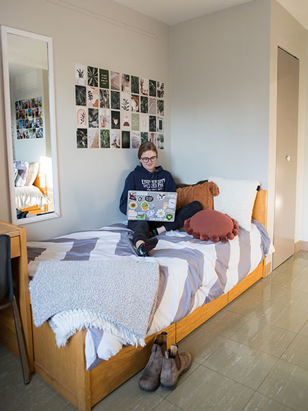 Female student sits on single bed in corner by poster wall, wooden desk at foot of bed
