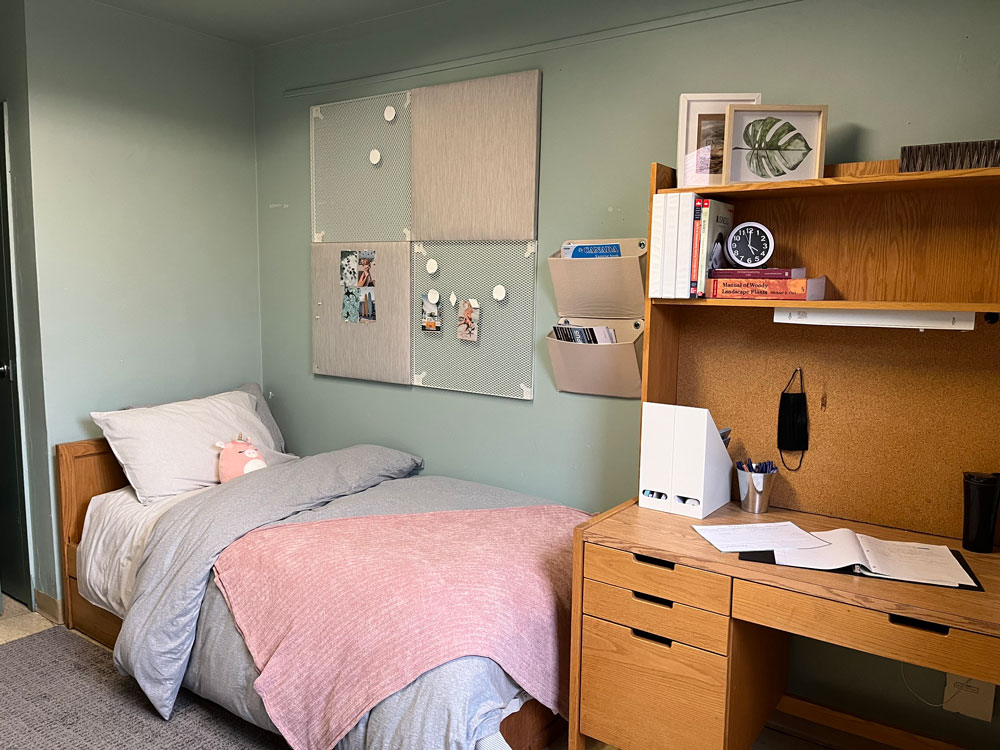 Single bed in corner with pink and grey blankets wooden desk to the right with pictures and books