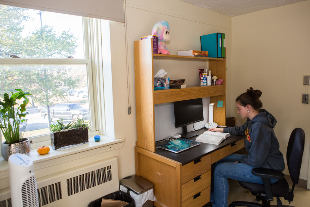 Student sits at a desk reading beside a large window with view of trees and parking lot