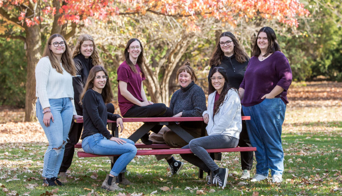 Group of female students standing beside and sitting on picnic table with fall trees behind them